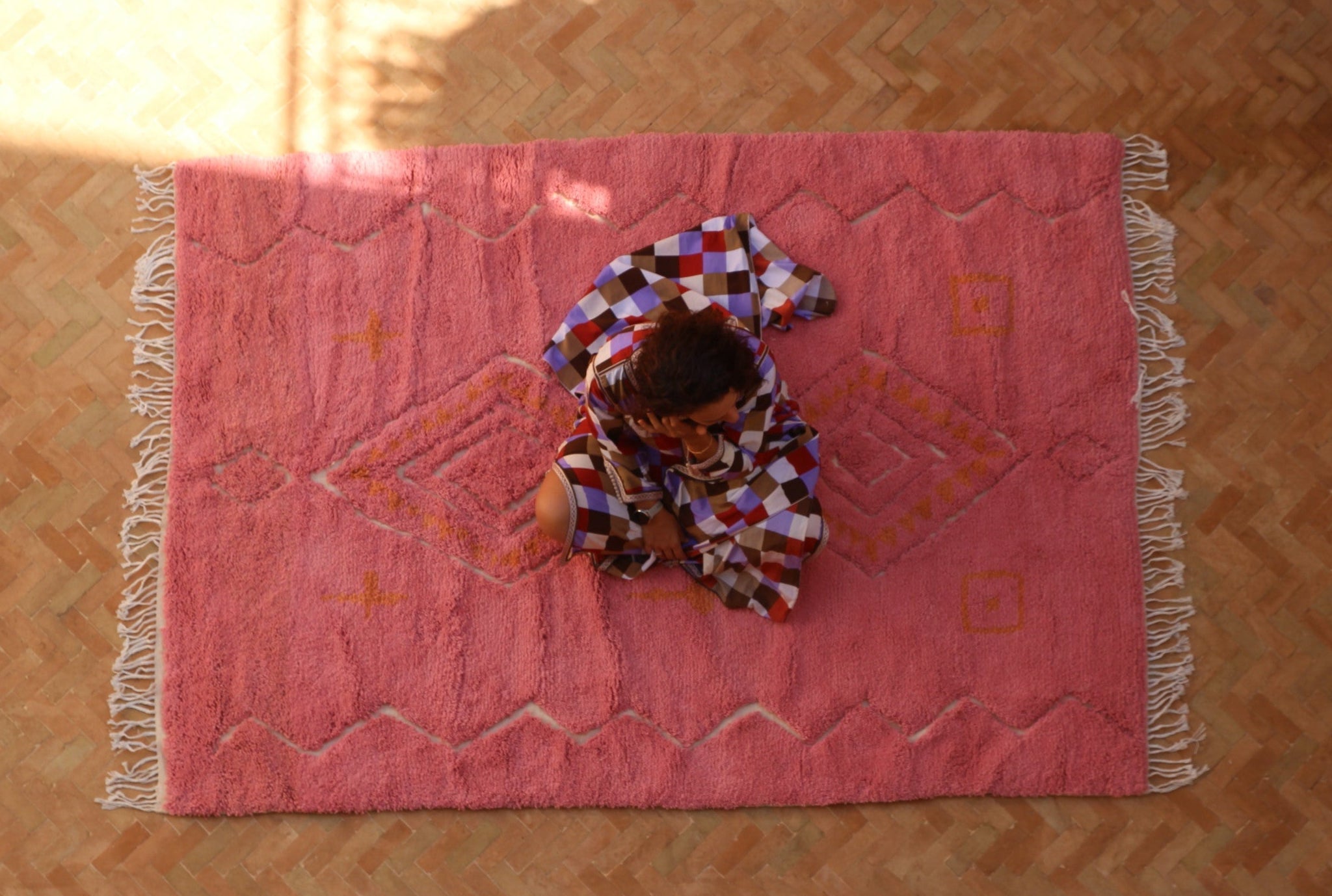 Person sitting on a pink patterned rug in a room with sunlight filtering through a window.