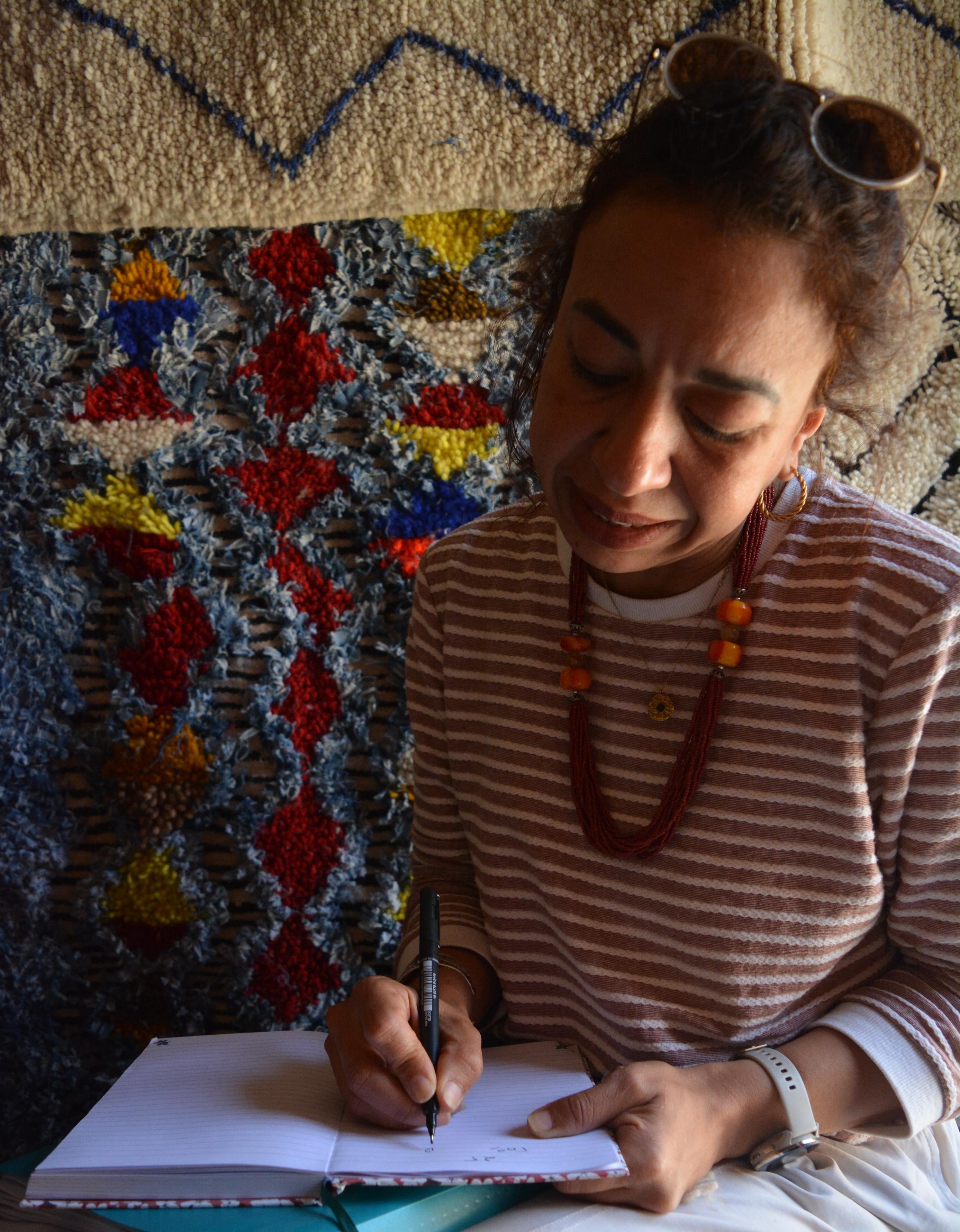 Woman sitting in front of a colorful rugs, writing in a notebook.